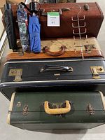 Four vintage suitcases stacked on top of each other with three umbrellas hanging and a metal belt hanger leaning against them.