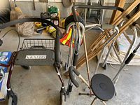View of the Dolomite walker, cane with folding seat, and other walking aids arranged on the floor of a garage space with tools and wooden items in the background.