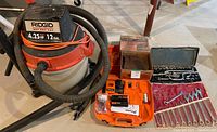 Photo of Rigid wet/dry vacuum with hose and attachments, Paslode battery charger with batteries, box of nails, socket set, and wrenches arranged on the floor
