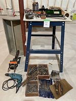 Workshop table setup with assorted tools and hardware, drill bits, Makita drill, Quick Vise clamp, and measuring instruments present on and under the table.