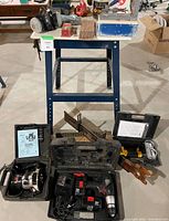 Workshop table with blue metal frame with multiple tools and cases on and under it, including Craftsman plunge router, Diehard cordless drill, Craftex staple gun, mitre saw, Kreg jig, and small items.
