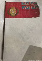 Red flag on wooden pole, vintage with heraldic crest and Union Jack, showing wear and holes.