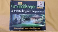 Front view of the box showing brand, product name, and image of a home garden area with sprinklers operating.