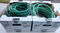 Two cardboard boxes each containing coiled green garden hoses, one hose with yellow section and metal fitting, the other hose has no visible fittings and shows usage wear.
