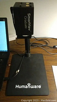 Photo showing SmartView Graduate video magnifier attached to an adjustable vertical stand with a branded base saying 'Humanware'. USB cable visible connected to the device.