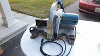 Full view of mitre saw showing blade, motor, handle, base, and power cord on a white table outdoors.
