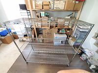 Full view of the metal shelving unit showing three wire shelves, round posts, and wheels for mobility. Surrounding space includes cardboard boxes and other storage items.