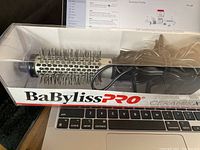 Photo shows the BaBylissPRO Ceramic Hot Air Styler in its packaging. The item is a cylindrical hot air brush with black bristles protruding from the ceramic barrel. The power cord is visible inside the box. The packaging is clear plastic, branded with BaBylissPRO CeramicX logos.