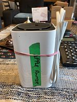 Front and side view of Apple AirPort Extreme router showing white body and attached power cable secured with a rubber band.