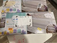 Two boxed Regalo swing down bed rails stacked on the floor, showing front and side views with clear product labels and descriptions.