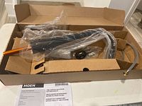 Box opened showing the kitchen faucet wrapped in plastic inside a brown box with Moen branding and model details visible on the packaging.