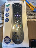 Photo showing the Philips branded universal remote control inside transparent packaging on a cardboard backing. Buttons and layout are clearly visible.