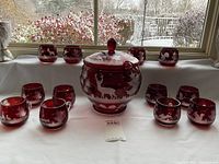 Full view of ruby red punch bowl at center with domed lid and twelve matching glasses arranged around it on a table by a window