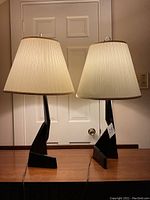 Two identical black zig zag base lamps with pleated off-white shades shown side by side on wooden surface, front view.