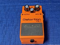 Top view of Boss DS-1 pedal showing orange body, rubber footswitch pad with Boss logo, three knobs and text labels.