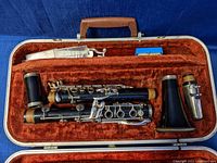 Open clarinet case showing disassembled clarinet parts including main body, bell, barrel, mouthpiece, ligature, reed case, and cleaning tools, inside brown velvet lined case.