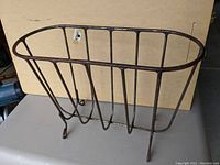 Steel magazine caddy shown at an angle revealing its curved legs, vertical steel bars, and an oval top rim.