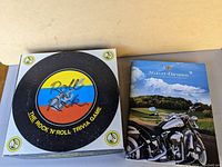 Overview of Rock n' Roll Trivia game box with round, colorful record design and Harley-Davidson catalog showing motorcycle on road and scenic background.