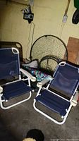 Two blue folding beach chairs with white metal frames, a round glass-top outdoor table, and a folding beach umbrella stand visible behind.