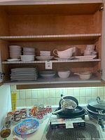 Wide cabinet shot showing stacks of white ceramic bowls, platters, and plates in upper shelves, along with colorful drinking glasses and kitchen items on the counter below