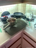 Photo showing various pots and pans arranged on a kitchen counter, including two pots with lids, a metal pot without lid, sauté pans, and a glass lid.