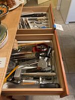 Two open wooden drawers containing assorted kitchen utensils including forks, spoons, mallets, spatulas, mashers, openers, basters, thermometers, and knives with wooden handles.