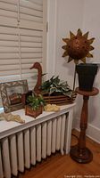 Wide view showing metal sun piece on stand, wooden goose planter with faux plant, framed mirror leaning, and assorted decorative items on radiator cover.