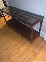 Full side perspective of bamboo framed console table with smoked glass top and rattan lower shelf on a hardwood floor.