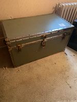 Front view of rectangular green wood trunk with metal borders, metal latches, and rubber handles, placed on carpeted floor near a radiator.