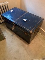 Top and angled front view showing overall metal storage trunk with locking clasp and rubber handle.