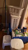 Photo of two white plastic laundry baskets stacked, white step stool, white waste bin with lid, blue plastic tote full of cleaning supplies including Swiffer and detergent.