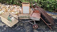 Photo showing vintage wooden Coca Cola crate, Economy wooden washboard, metal wagon with wooden bed, and wooden sled on asphalt ground near greenery