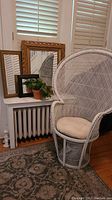 White wicker chair with removable beige cushion next to a radiator topped with three framed mirrors and a clay planter with faux succulent.