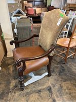 Side view of wood armchair showing leather seat, upholstered back with metal stud trim, and carved wood arm with scratches.