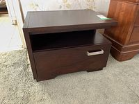 Front and side view of dark wood bedside table showing drawer with metal pull and open shelf underneath.