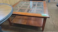 Front and side view of the square coffee table showing the wood frame, glass top with grid pattern, and lower wooden shelf.