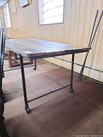 Side angle of dining table showing wood plank top and black pipe metal base with caster wheels.