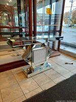 Front angle view of a mirrored hall table positioned in front of a large glass cabinet and window, showing the octagonal mirrored base and rectangular bevelled mirrored top reflecting the tiles and surroundings.