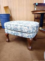 Front-side view of the floral upholstered ottoman showing the detailed fabric pattern and wooden legs.