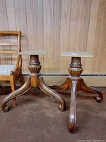 Pair of wooden table pedestals with visible carved details and curved legs under square wooden tops, placed against wood paneled wall next to a chair.