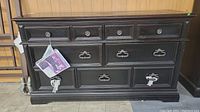Front view of black Pulaski dresser with multiple drawers and oversized pulls, featuring a tag attached to one pull