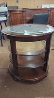 Side view of round wood side table with glass top and cane shelf below.