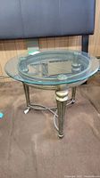 Front angle showing round glass top and metal decorative frame with braided edge and tapered legs.