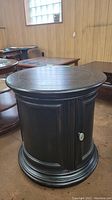 Round dark wood side table with a closed door, showing the overall shape and style.
