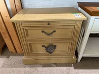 Front view of beige Stanley bedside table showing two drawers with ornate metal pulls and a small top drawer knob.