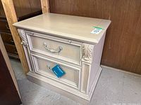 Front angled view showing the bedside table's two drawers, decorative carved accents, metal pulls, and cream finish.