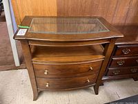 Front view of wooden bedside table with glass inset top, two drawers with metal pulls, and lower shelf.