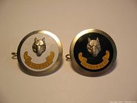 Front view of two round Boy Scouts Wolf Cub cap badges, one with white background, one with black background, featuring wolf head and banner.