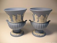 Front view of two Wedgwood mini urn vases, showing different classical white relief designs on round blue jasperware bases with fluted lower portions.