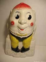 Front view of ceramic Humpty Dumpty bank showing detailed facial features and painted attire, including a red hat and yellow pants.